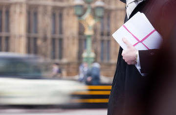 A barrister in front of a road and building carrying files tied in a pink ribbon.
