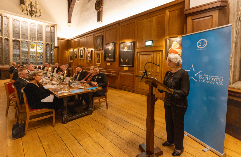 A photo of a woman speaking at a podium with a dinner table of people watching her in the background